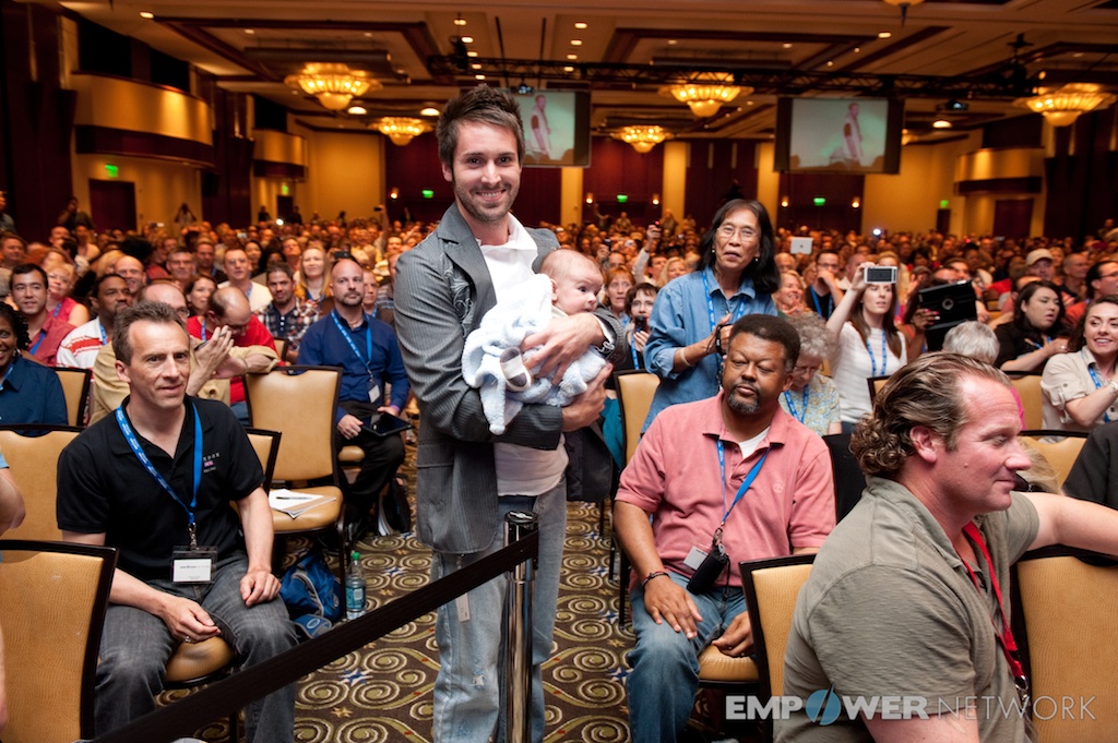 Smiling attendee cradling a baby while standing in a packed audience at Empower Network's 2012 Atlanta event